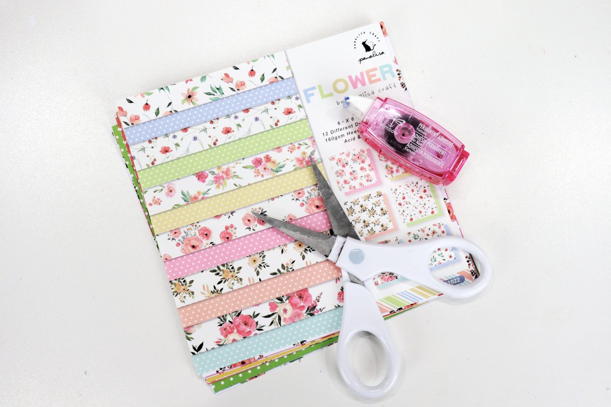 Image contains a stack of patterned scrapbook paper, a pink adhesive tape runner, and a pair of white handled scissors on a white background.