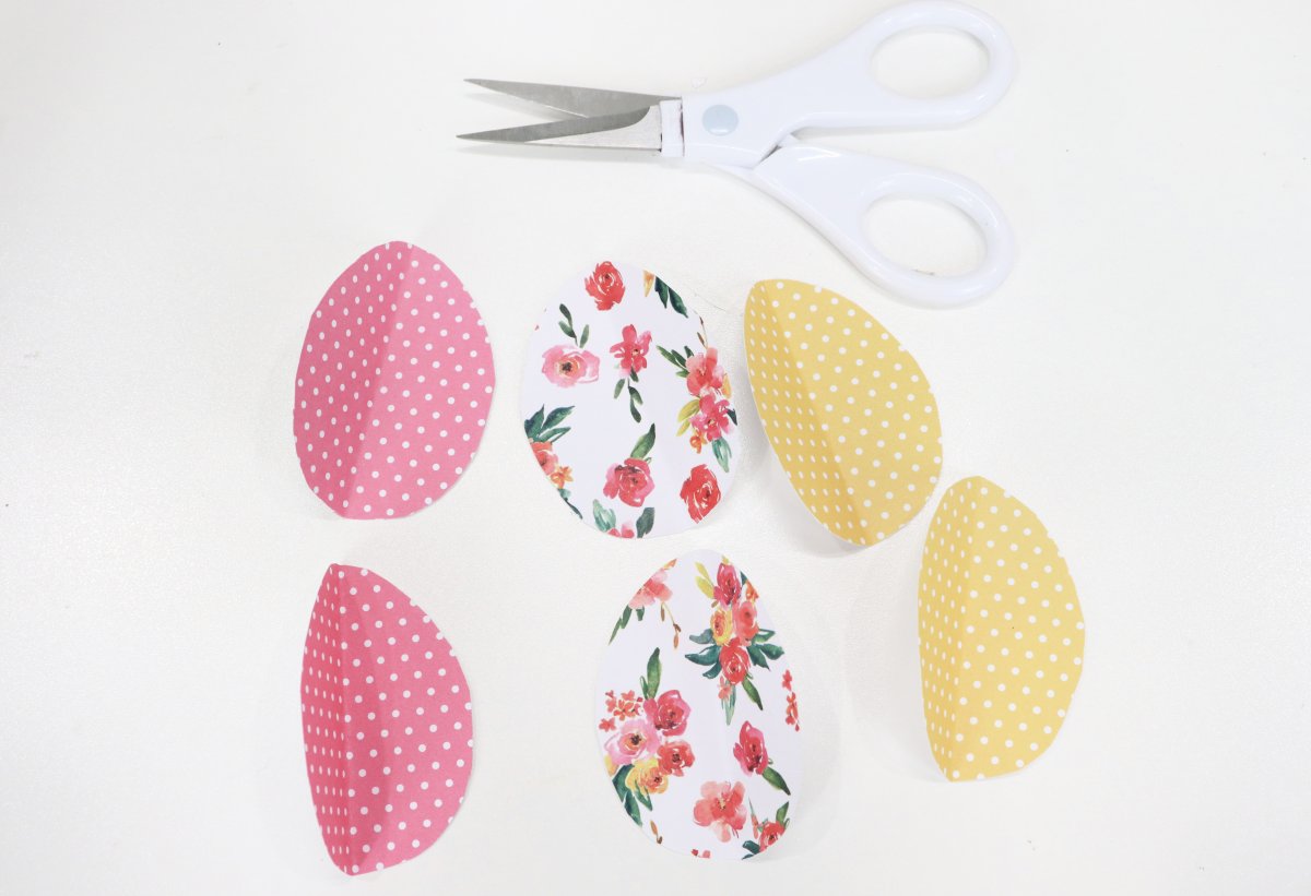 Image contains six paper egg shapes cut from patterned scrapbook paper in pink, white, and yellow. They sit on a white background with a pair of white-handled scissors above.