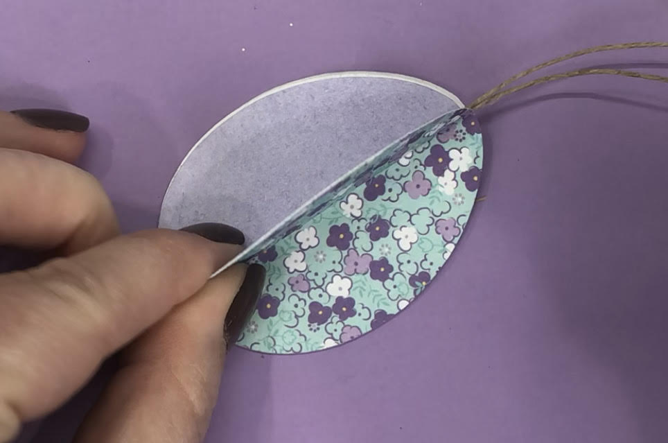 Image contains a hand adhering a 3-D paper egg together. The egg is made of purple and teal patterned scrapbook paper and is on a purple background.