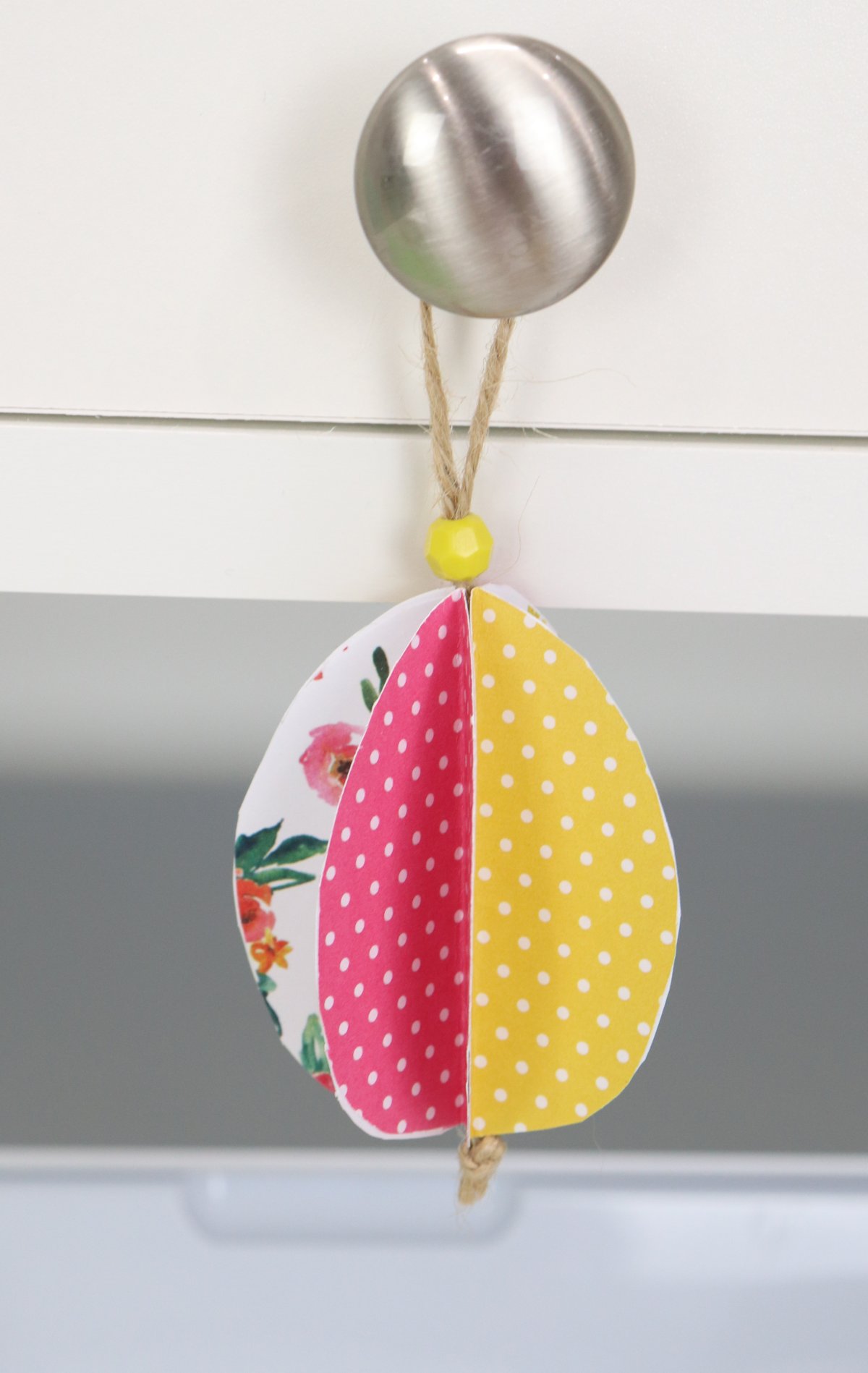 Image contains a 3-D paper egg made of patterned scrapbook paper hanging from a silver drawer knob. 