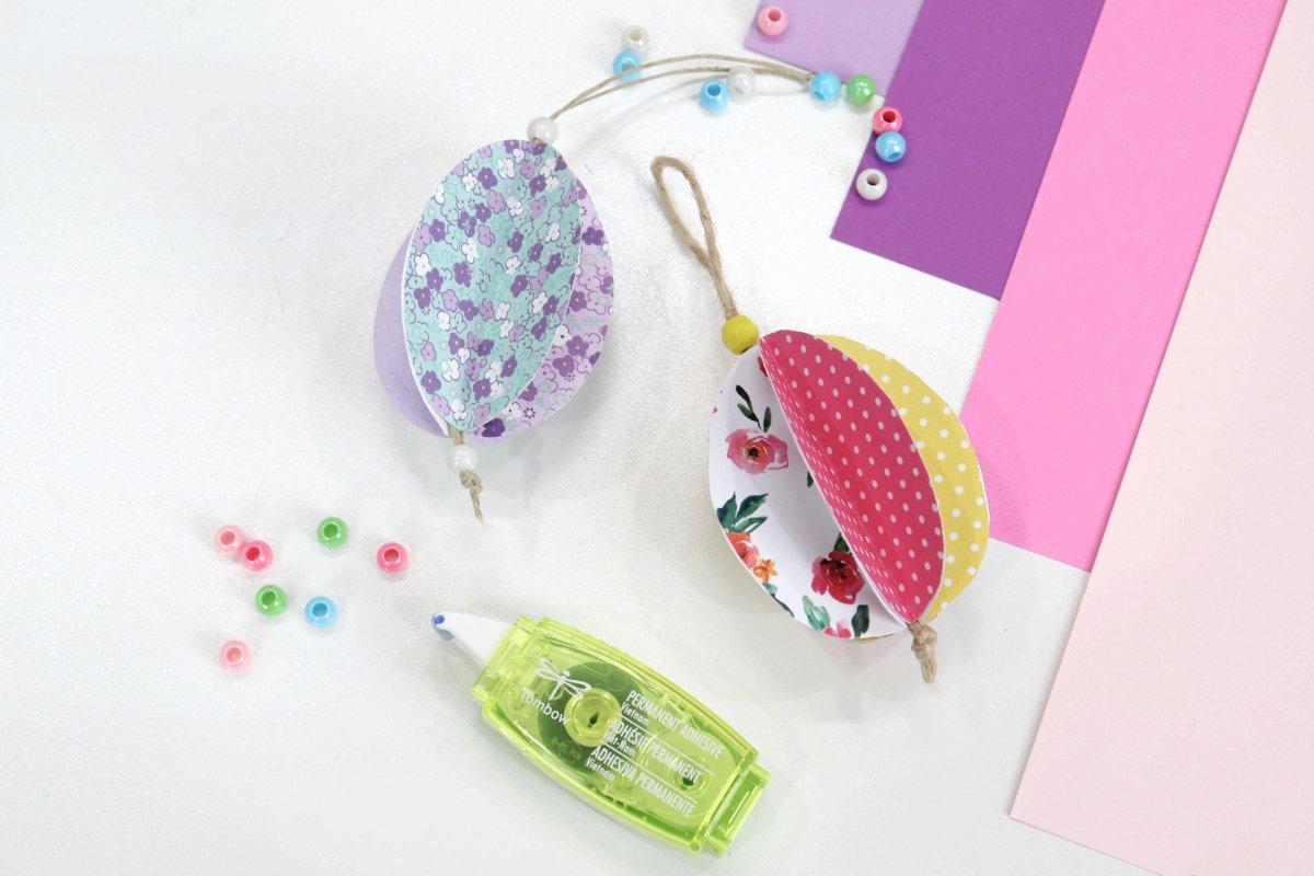 Image contains two 3-D paper eggs made of patterned scrapbook paper in teal, purple, white, pink, and yellow. They sit on a white background, surrounded by multi-colored beads, pink and purple cardstock, and a right green adhesive tape runner.