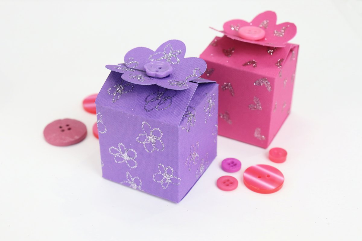 Image contains two boxes made of folded cardstock; one purple and one pink. They are on a white background with pink buttons scattered around.