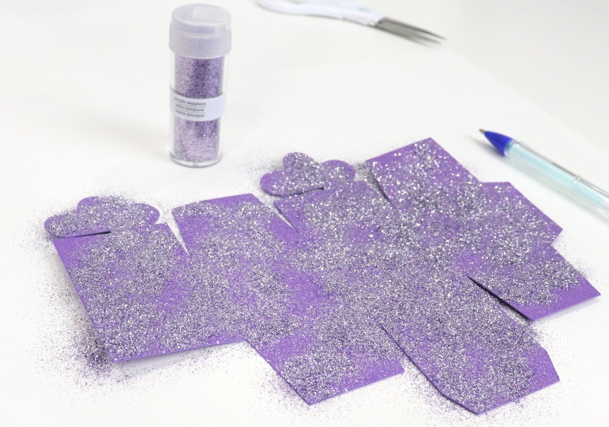 Image contains a tube of fine purple glitter, a glue pen, a pair of small white-handled scissors, and a piece of purple cardstock covered in the fine purple glitter, all on a white background.