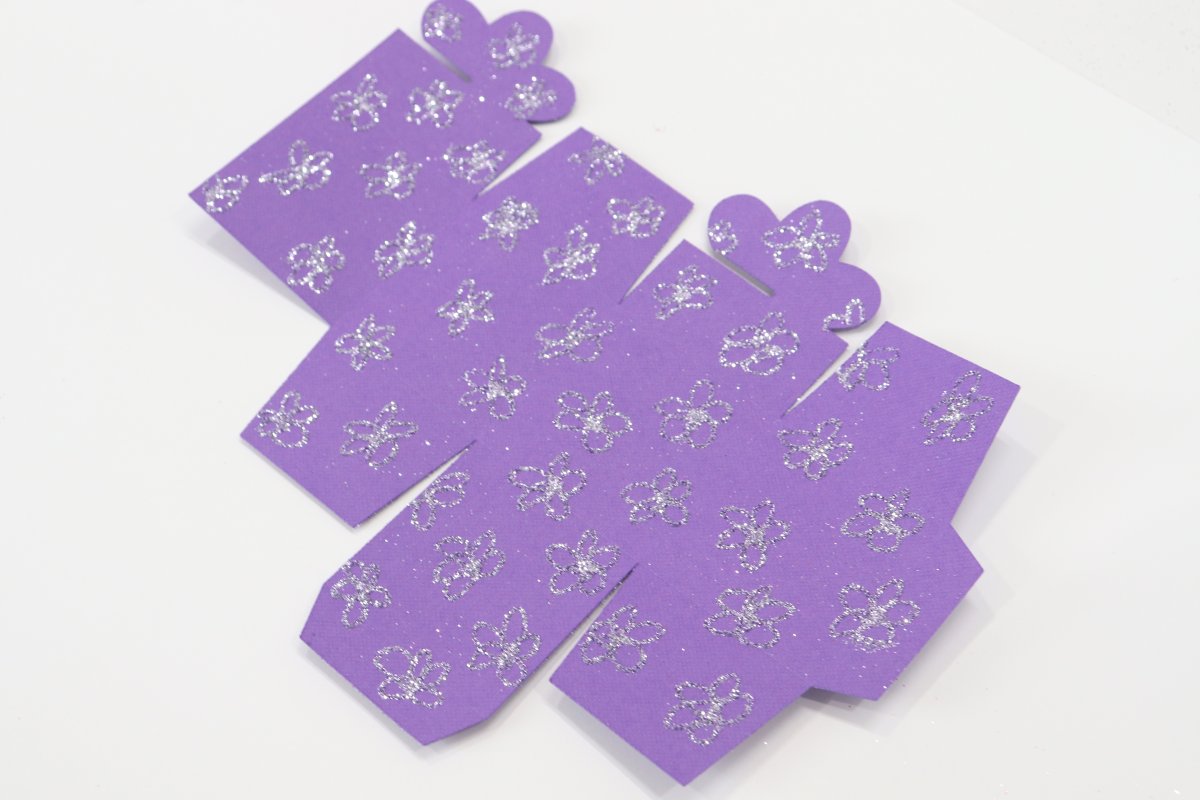 Image contains a piece of purple cardstock cut into the shape of a box template, with a pattern of flowers all over it in light purple glitter.