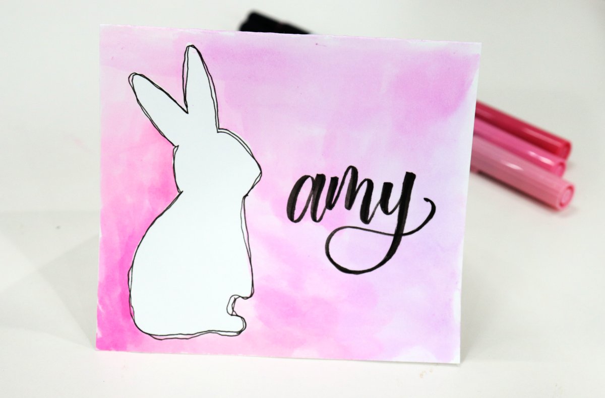 Image contains a placecard featuring a white bunny outlined in black on a pink and purple watercolor background with the name, "amy" written in black.
