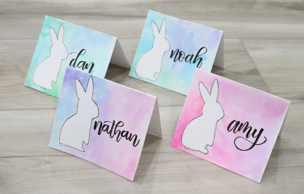 Image contains four placecards, each featuring a white bunny on a multi-colored watercolor background and a name written in black marker.
