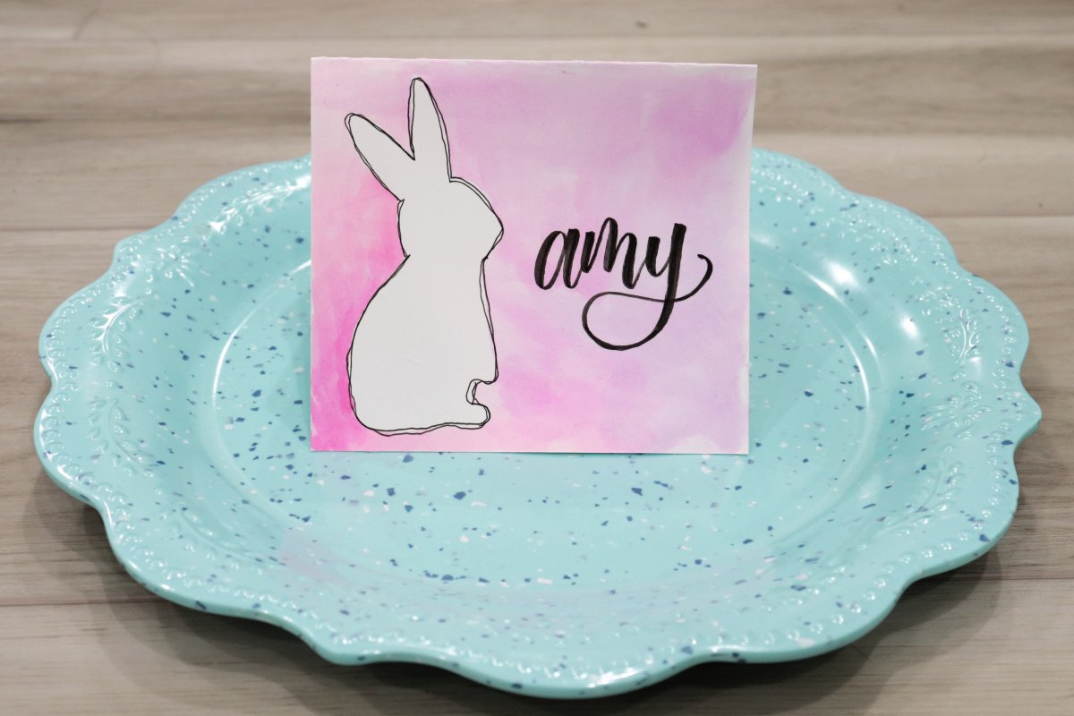 Image contains a teal plate with a scalloped edge on a wooden surface. On the plate is a placecard featuring a white bunny outlined in black on a pink and purple watercolored background. Next to the bunny, the name "amy" is written in black.