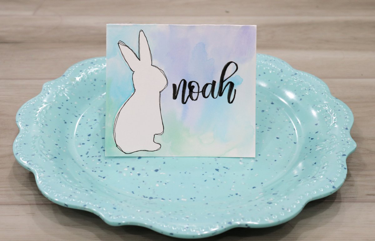 Image contains a teal speckled plate with scalloped edges sitting on a wooden surface. On the plate is a placecard featuring a white bunny on watercolor background of mixed blues, greens, and purples, and the name "Noah."