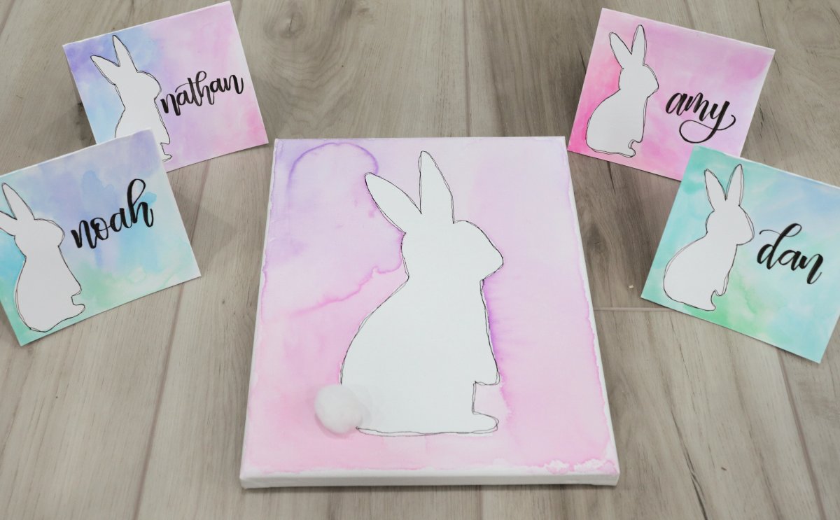 Image contains a canvas and four placecards, each with a white bunny on a multi-colored watercolor background, sitting on a wooden surface.