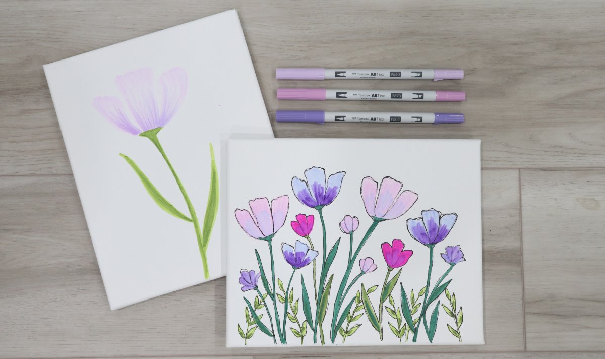 Image contains two white canvases decorated with hand-drawn pink and purple flowers. They sit on a wooden background, along with three purple markers.