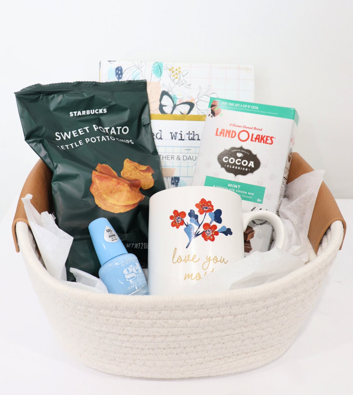 Image contains a cream colored basket holding a bag of sweet potato chips, a mother-daughter journal, a package of mint cocoa, a bottle of blue nail polish, and a coffee mug, on a white background.