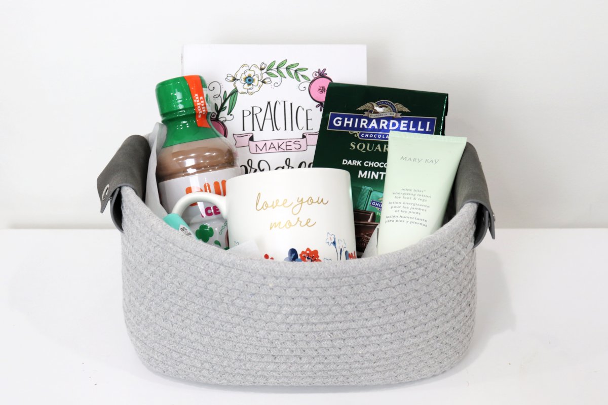 Image contains a grey basket holding a journal, a bottled coffee drink, a package of Ghirardelli chocolates, a bottle of nail polish, a coffee mug, and a tube of lotion, on a white background.
