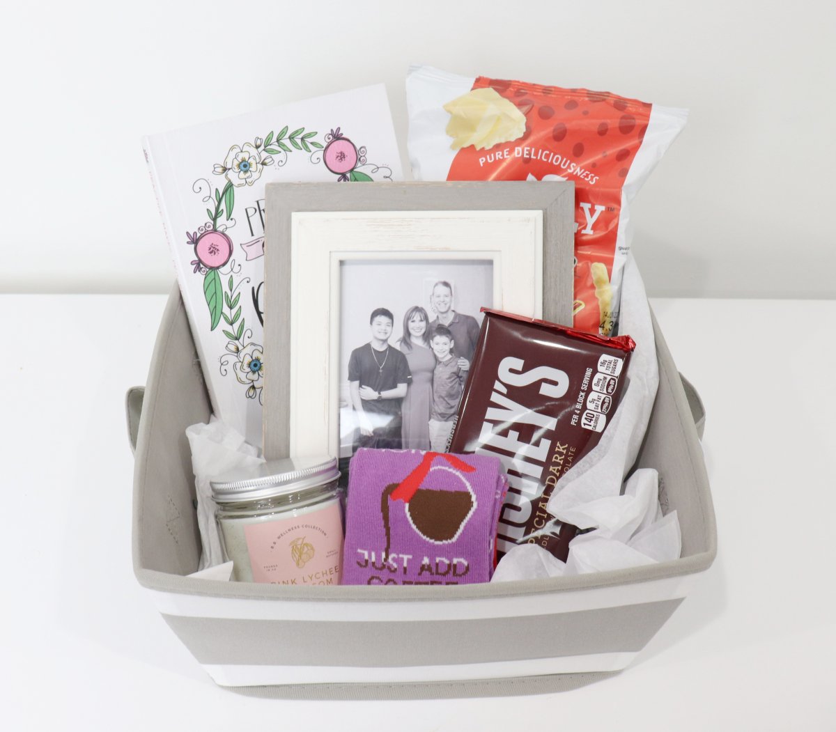 Image contains a grey and white striped basket holding a journal, a bag of cheese curls, a framed black and white photo of Amy's family, a special dark Hershey bar, a jar candle, and a pair of purple socks, on a white background.