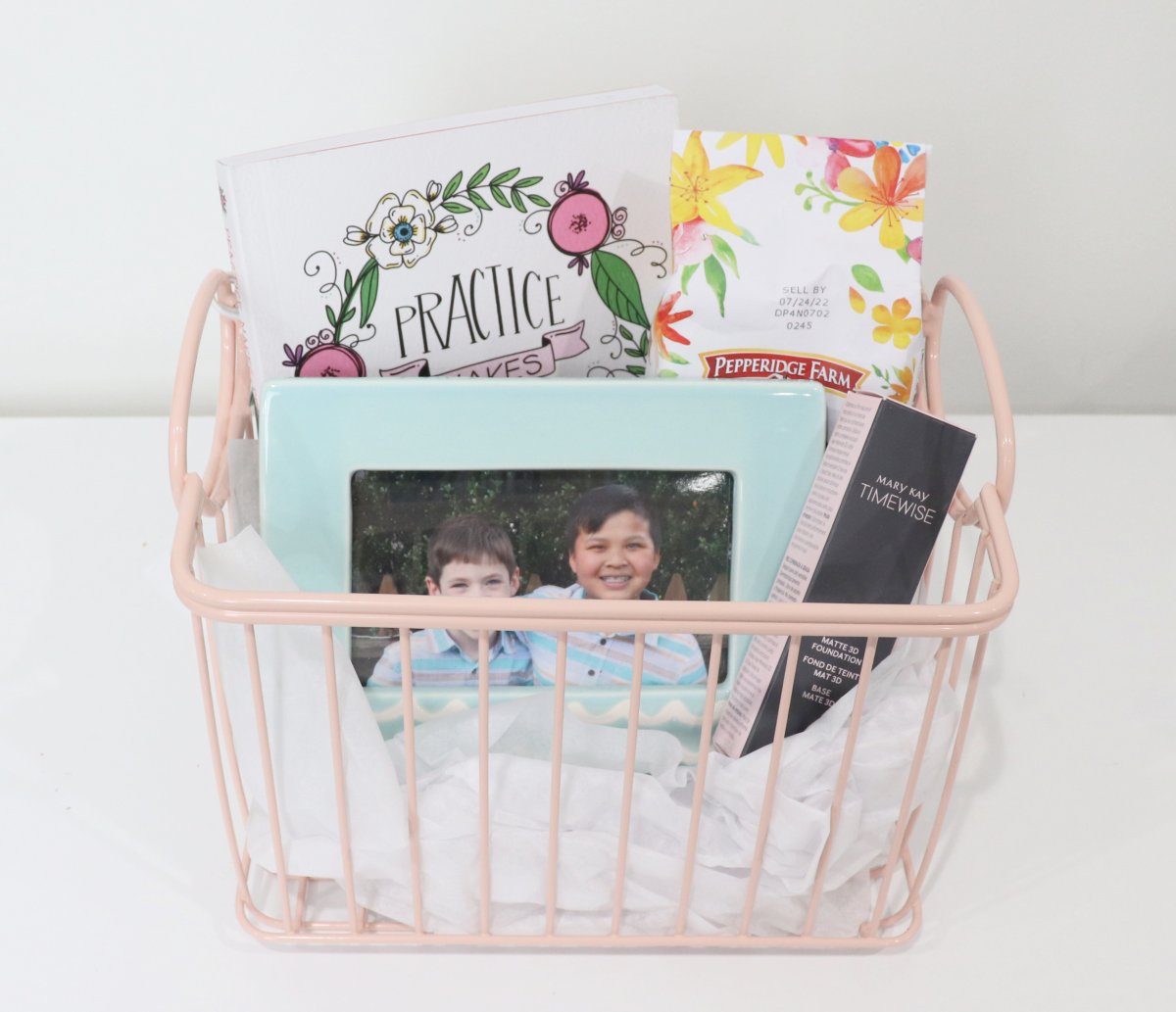 Image contains a pink wire basket holding a journal, a package of cookies, a framed photo of two boys, and a tube of foundation, on a white background.