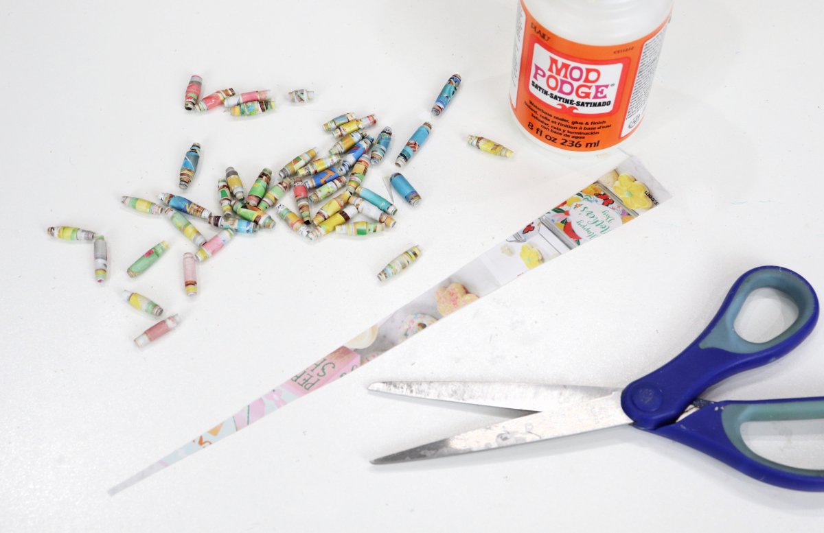 Image contains an assortment of paper beads, a triangle cut from a magazine page, a jar of Mod Podge, and a pair of blue handled scissors on a white background.