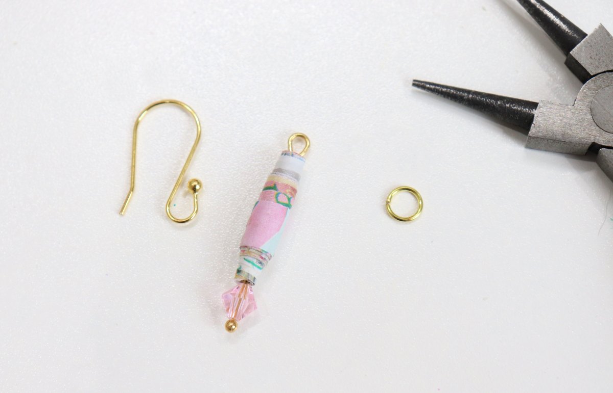 Image contains a rolled paper bead and a pink crystal bead on a gold head pin, next to a gold ear wire, a gold jump ring, and a pair of pliers on a white background.