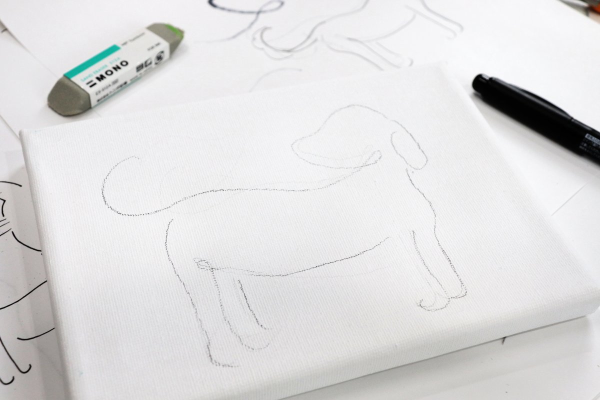 Image contains a white canvas with a pencil sketch of the outline of a dog. It sits on a background of sketch paper covered with similar images, and a marker and eraser sit nearby.