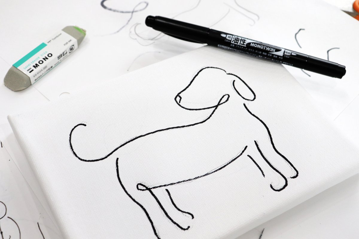 Image contains a white canvas with an image of a black line art dog. A marker and eraser sit nearby and the background is paper with sketches of the image in pencil and black pen.