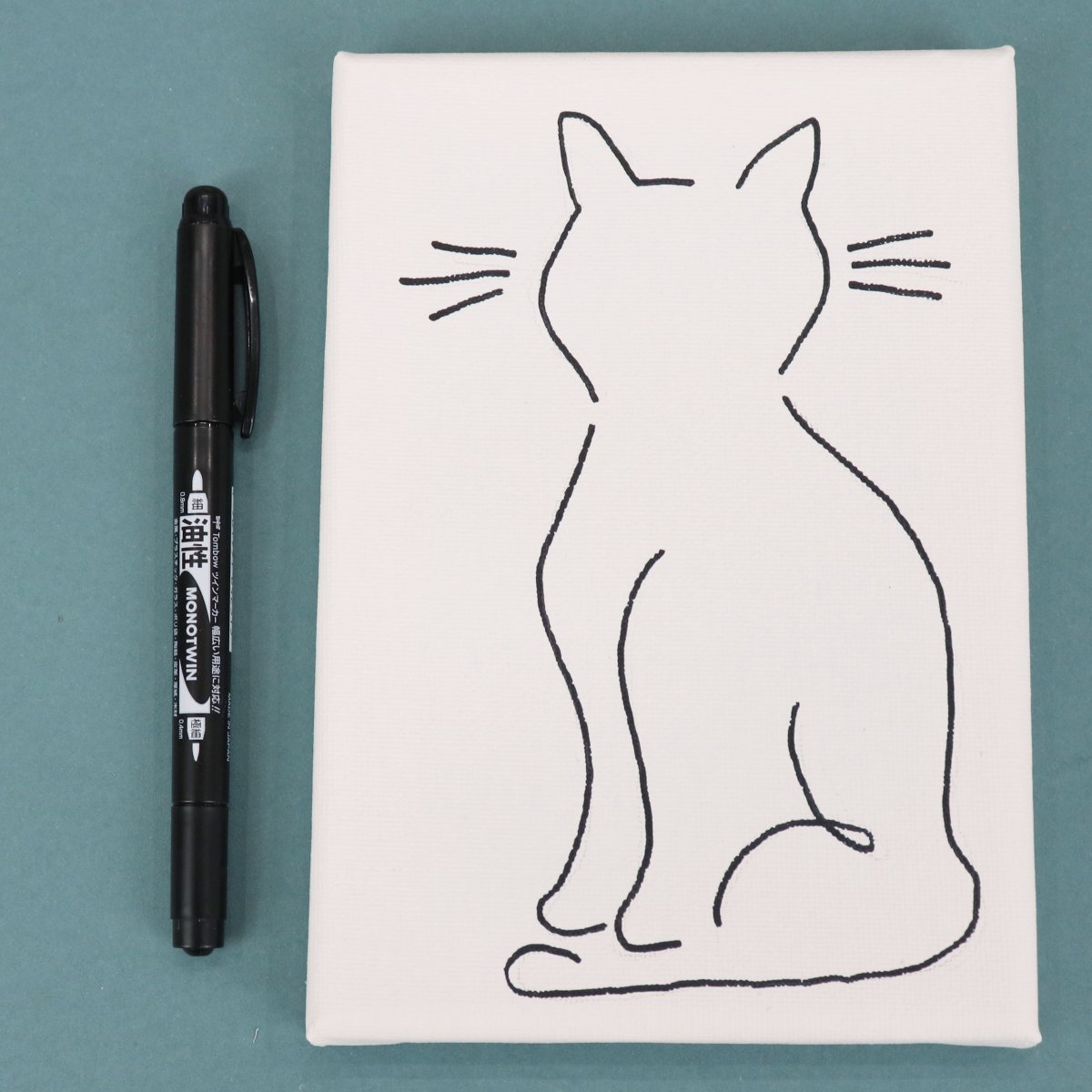 Image contains a white canvas with a line drawing of a seated cat. It is on a teal background with a Tombow MONO Twin marker next to it.
