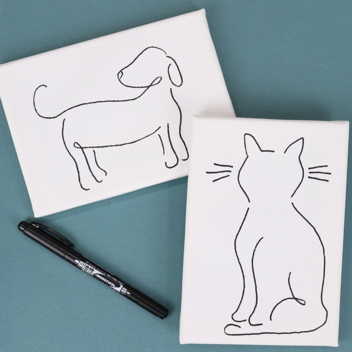 Image contains two white canvases; one with a minimalist line drawing of a dog on it, and the other with a line drawing of a cat. They sit on a teal background with a black marker nearby.