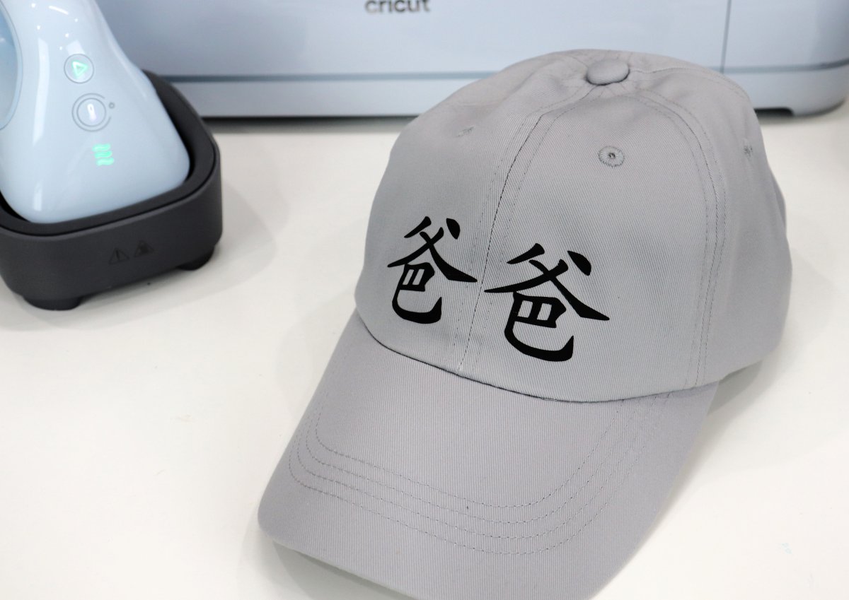 Image contains a grey ball cap with the Chinese characters "baba" on the front in black. The Cricut Hat Press and blue Cricut Maker 3 machine are in the background.