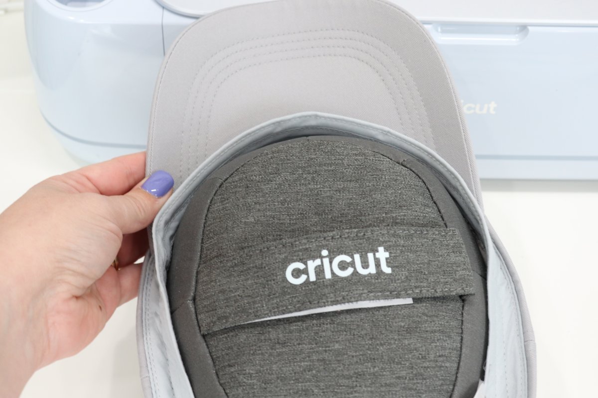 Image contains Amy's hand holding a grey ball cap stretched over the Cricut hat form. A blue Cricut Maker 3 is in the background.