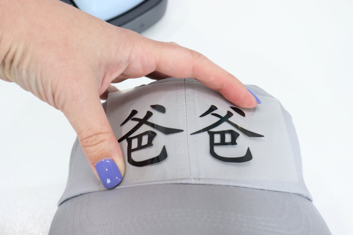 Image contains Amy's hand holding an iron-on design in place on a grey ball cap. The Cricut Hat Press is in the background.