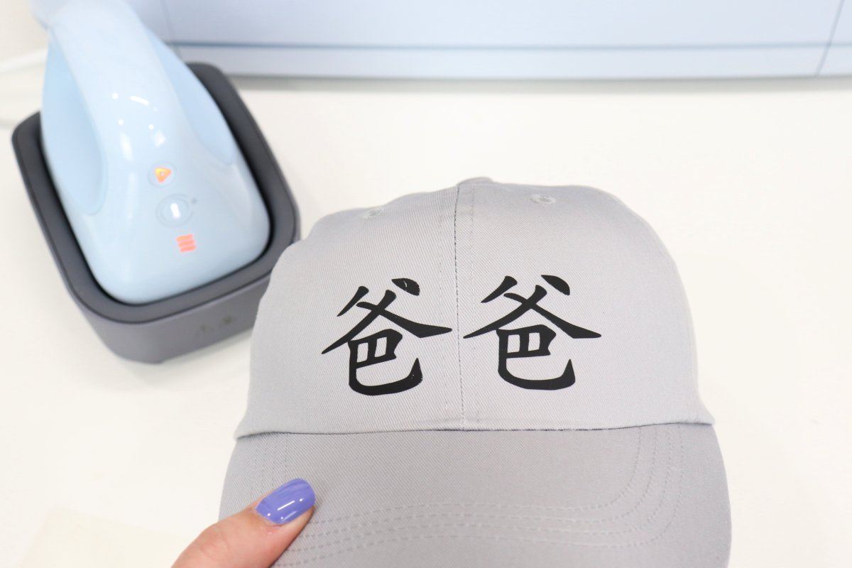 Image contains a grey ball cap with the Chinese characters "baba" on the front in black. The Cricut Hat Press and Cricut Maker 3 machine are in the background.