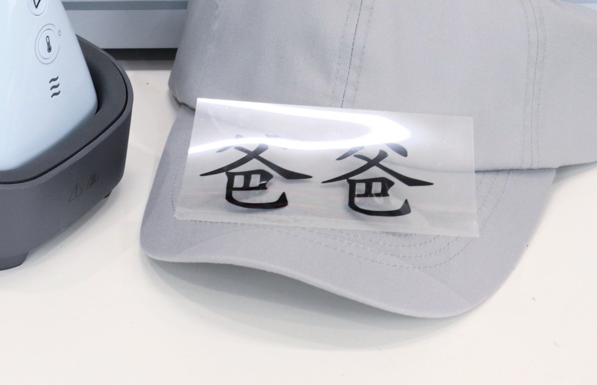 Image contains a grey ball cap, a blue Cricut Hat Press, and the Chinese characters "baba" cut from iron on black vinyl adhered to a clear carrier sheet.