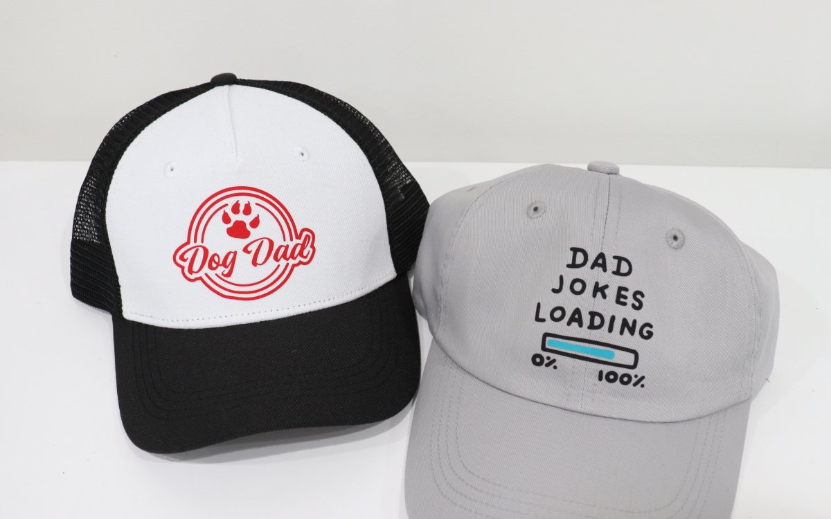 Image contains two hats; a black and white trucker hat with a red design that reads, "Dog Dad", and a grey ball cap that says, "Dad Jokes Loading" in black vinyl.