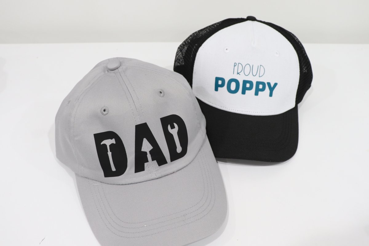 Image contains two hats; a grey ball cap with the word "DAD" in black iron-on vinyl, and a black and white trucker hat with the words "Proud Poppy" in teal.