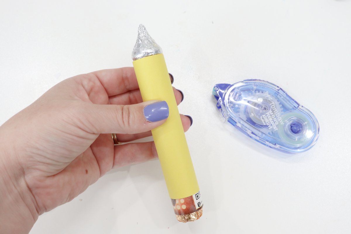 Image contains Amy's hand holding a roll of Rolos wrapped in yellow cardstock with a Hershey's Kiss on top, and a blue adhesive runner beside it on a white table.