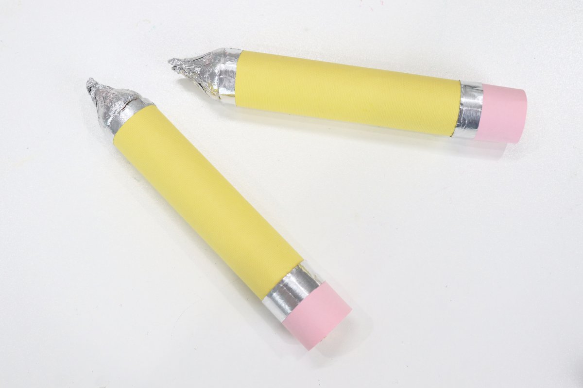Image contains two candy pencils made from Rolo candies wrapped in yellow and pink cardstock and foil, and Hershey's kisses.