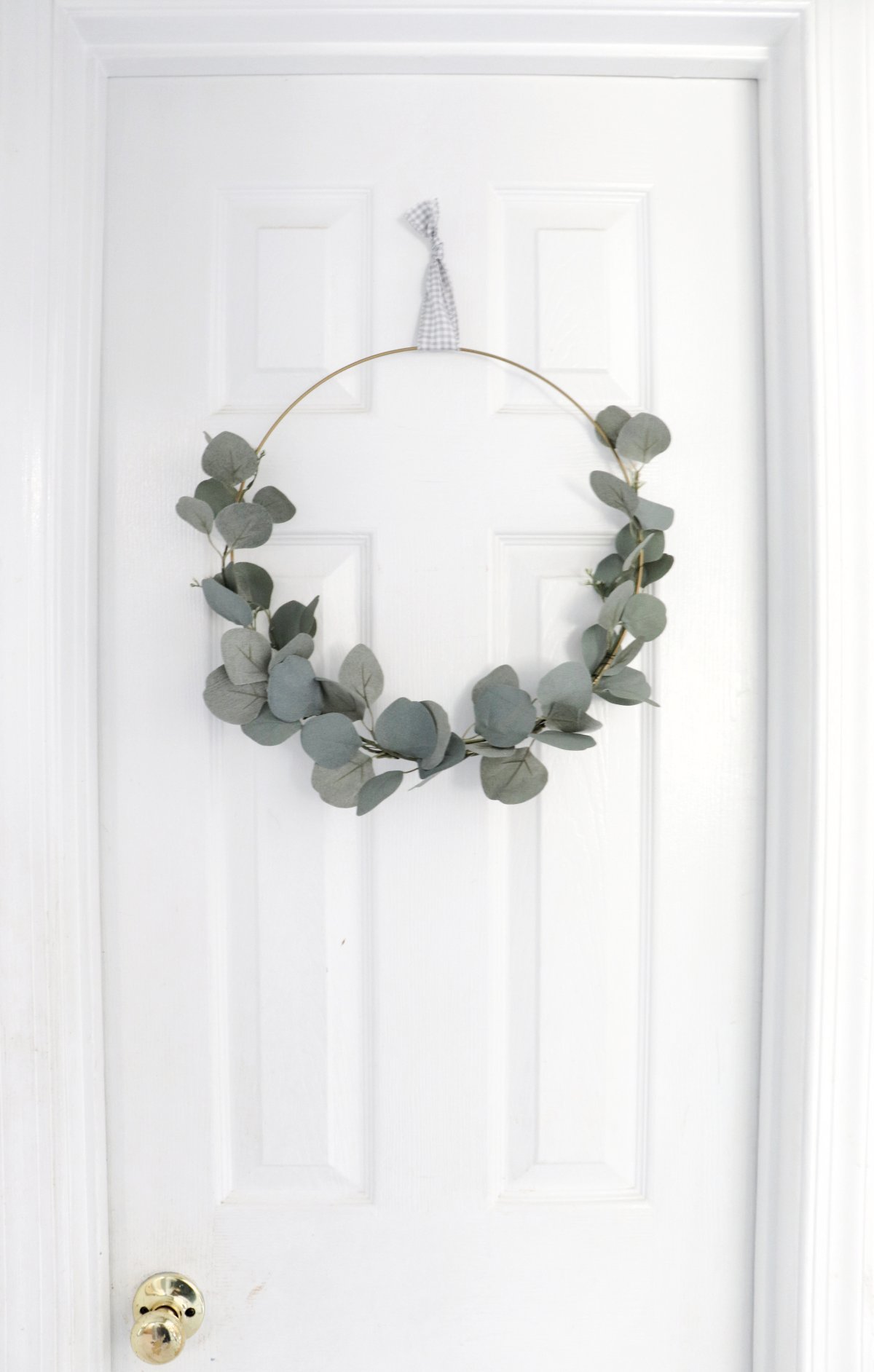 Image contains a faux eucalyptus hoop wreath hanging on a white door.
