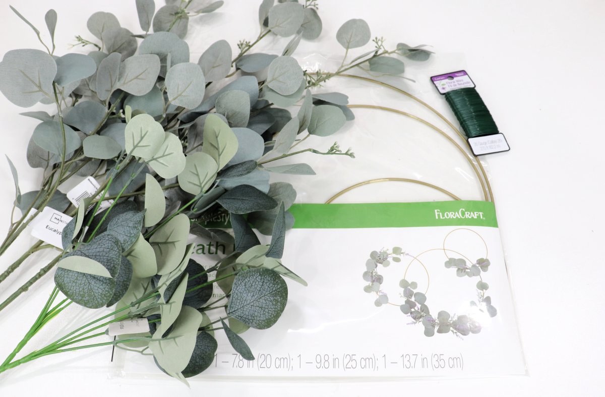 Image contains stems of faux eucalyptus, a pack of three gold wreath hoops, and a spool of green floral wire on a white background.