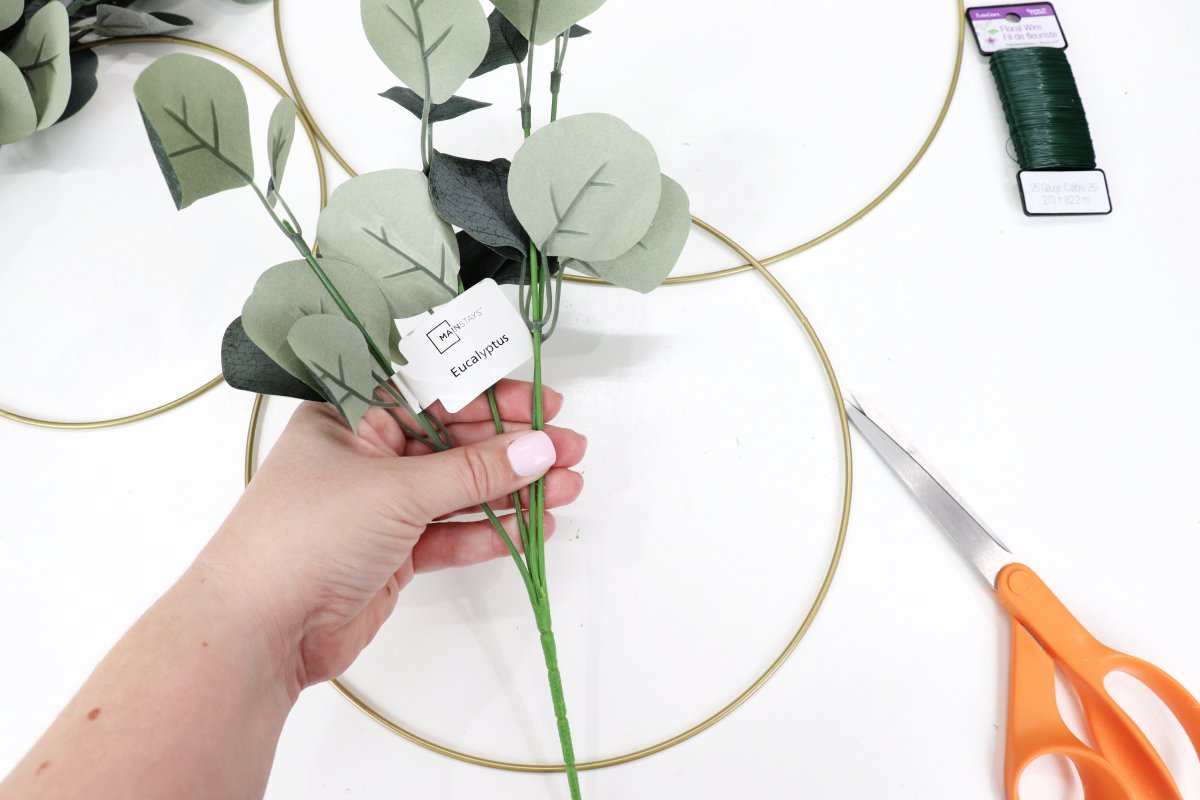 Image contains Amy's hand holding a pick of faux eucalyptus. In the background are three gold hoops, a pair of orange handled scissors, and a spool of floral wire.