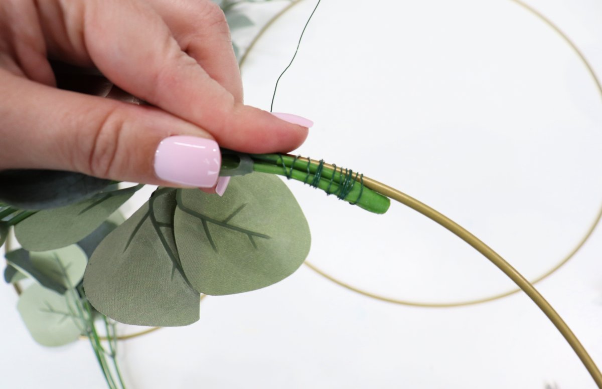 Image contains Amy's hand holding a stem of faux eucalyptus wired onto a gold hoop. Another gold hoop is in the background.
