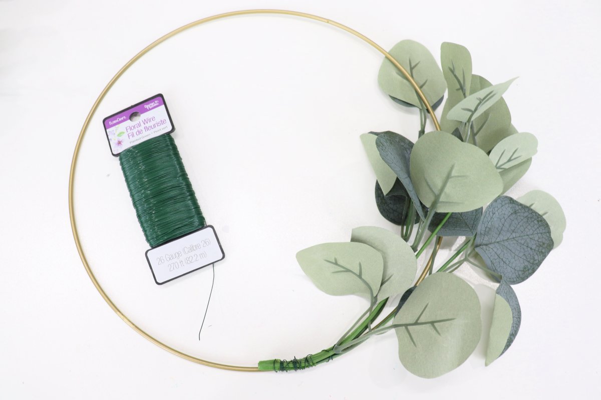 Image contains a gold hoop with a stem of faux eucalyptus wired to one side. A spool of floral wire sits inside the hoop on a white background.