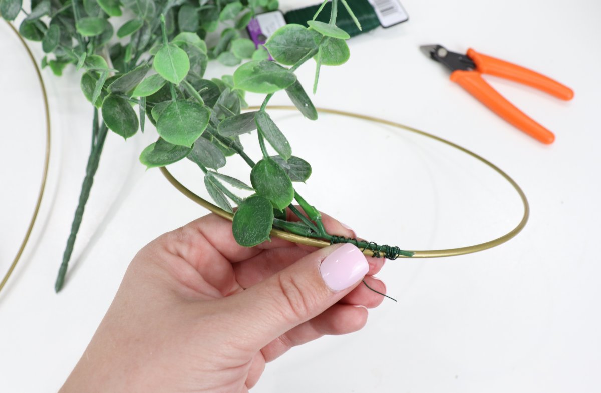 Image contains Amy's hand holding a gold wreath hoop with faux eucalyptus wired to it. A pair of orange handled pliers and a spool of floral wire are in the background. 