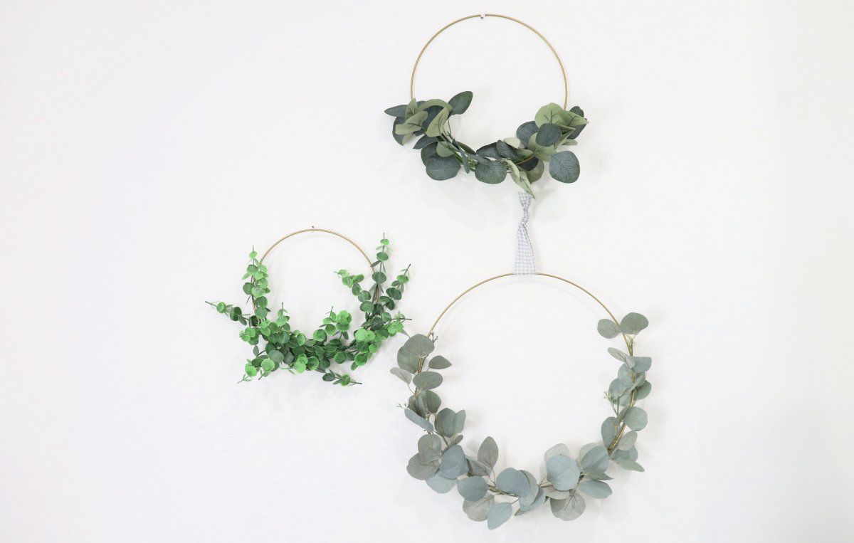 This image contains three wreaths; a small, medium, and large size. Each wreath is a gold hoop with the bottom half covered by faux eucalyptus. The wreaths hang on a white background.