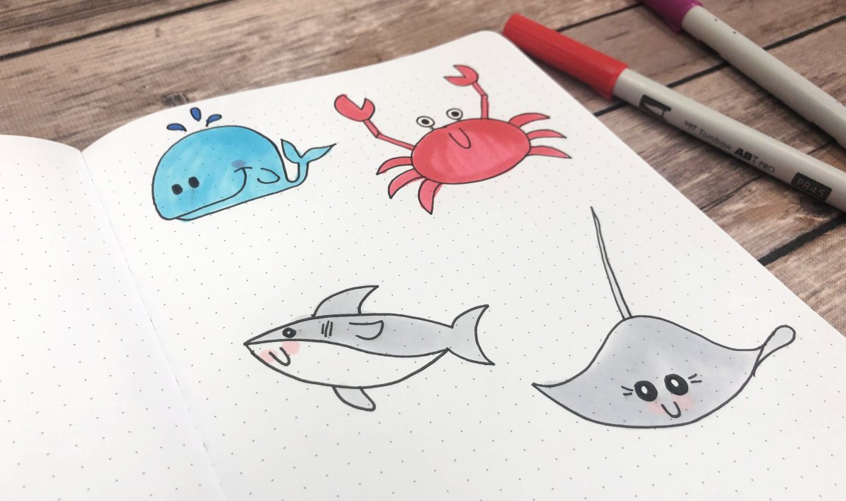 Image contains an open sketchbook with drawings of a whale, a crab, a shark, and a stingray. It sits on a wooden background with two markers to the side.