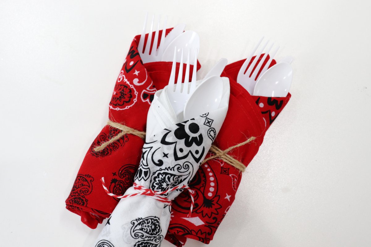 Image contains three sets of plastic utensils, each rolled in a red or white bandana and tied off with twine, on a white background.