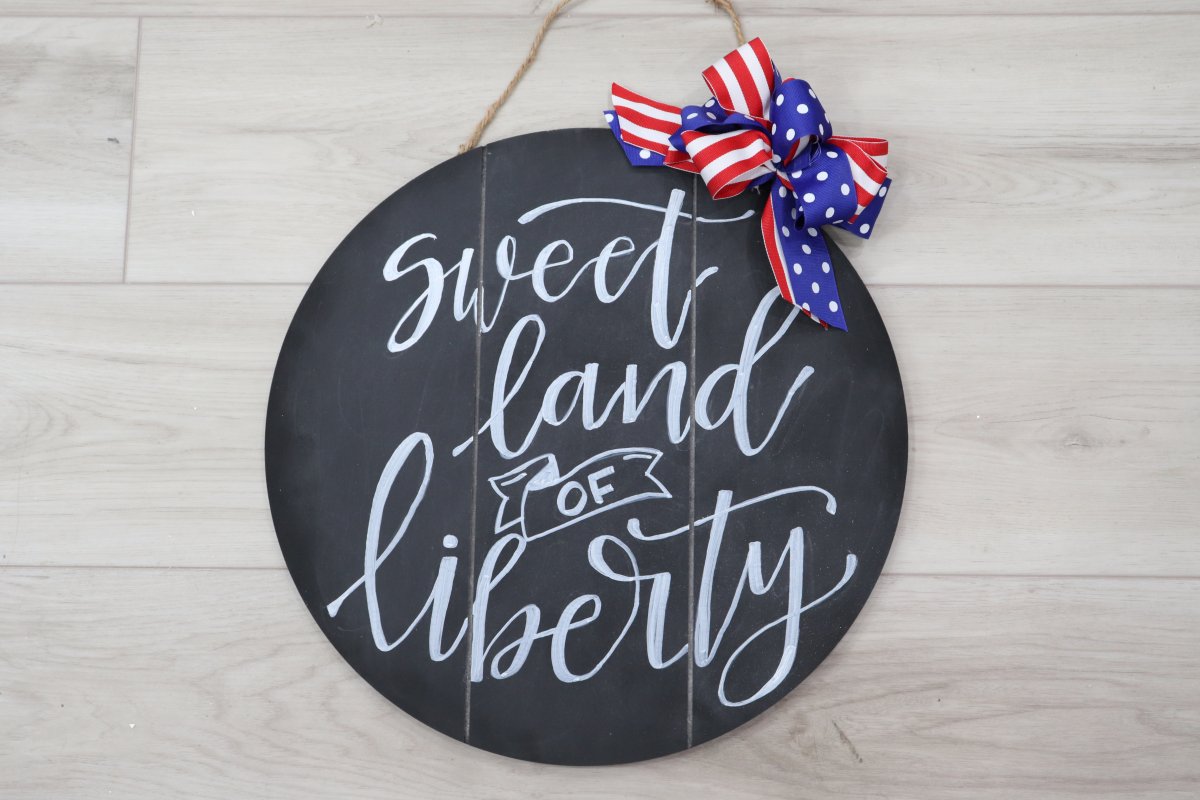 Image contains a round chalkboard sign with the words, "sweet land of liberty" written on it in white chalk marker, and a red, white, and blue ribbon bow on top.