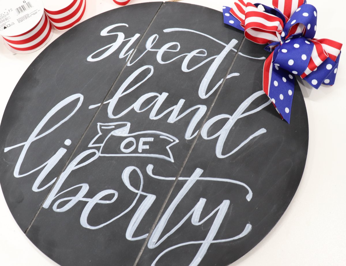 Image contains a round chalkboard sign with the words, "sweet land of liberty" in white chalk marker and a red, white, and blue bow.