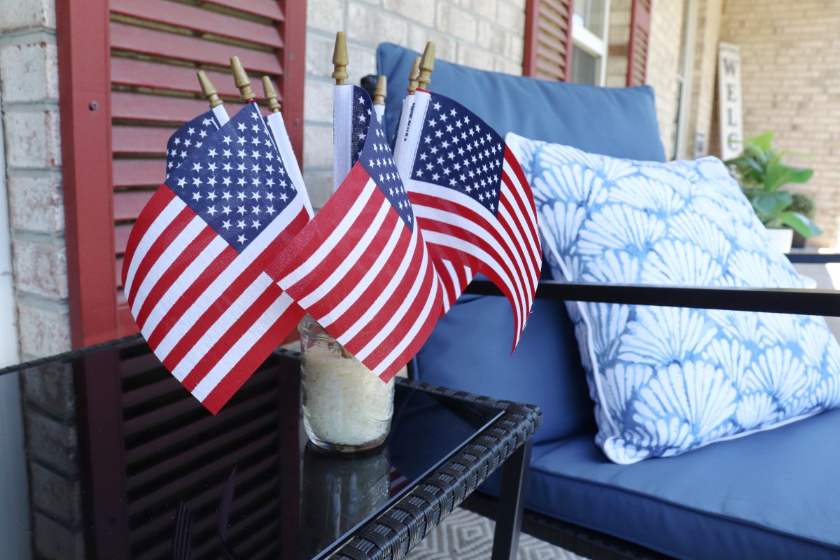 Image contains a mason jar filled with rice and 7 mini American flags. It sits on a black outdoor table. In the background is a front porch with a blue rocking char, a blue and white pillow, red shutters, a plant, and a welcome sign.