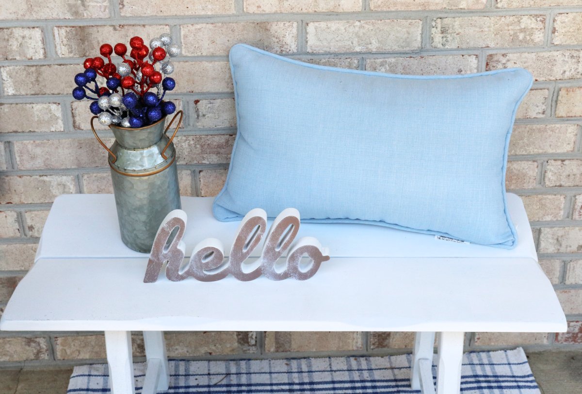 Image contains a white bench sitting against a brick wall with a blue and white plaid rug underneath. On the bench is a blue pillow, a cutout wooden "hello," and a galvanized tin milk jug filled with red, silver, and blue berries.