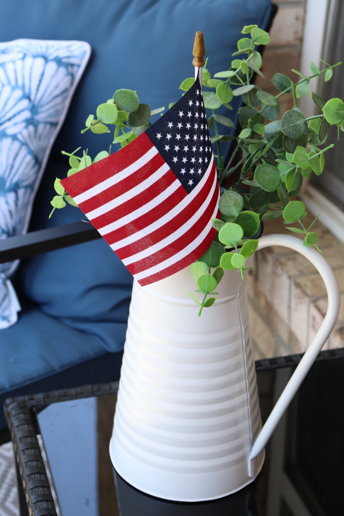 Image contains a white pitcher filled with faux eucalyptus and a small American flag. It sits on a black outdoor table with a blue outdoor rocking chair in the background.