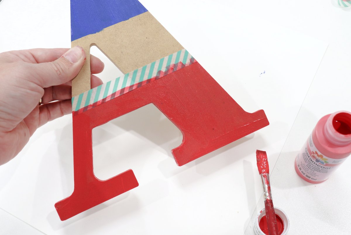 Image contains a wooden letter "A." The top third is painted blue, the bottom third is painted red and taped off with washi tape. A jar of red paint and a paintbrush sit on the table beside the letter.