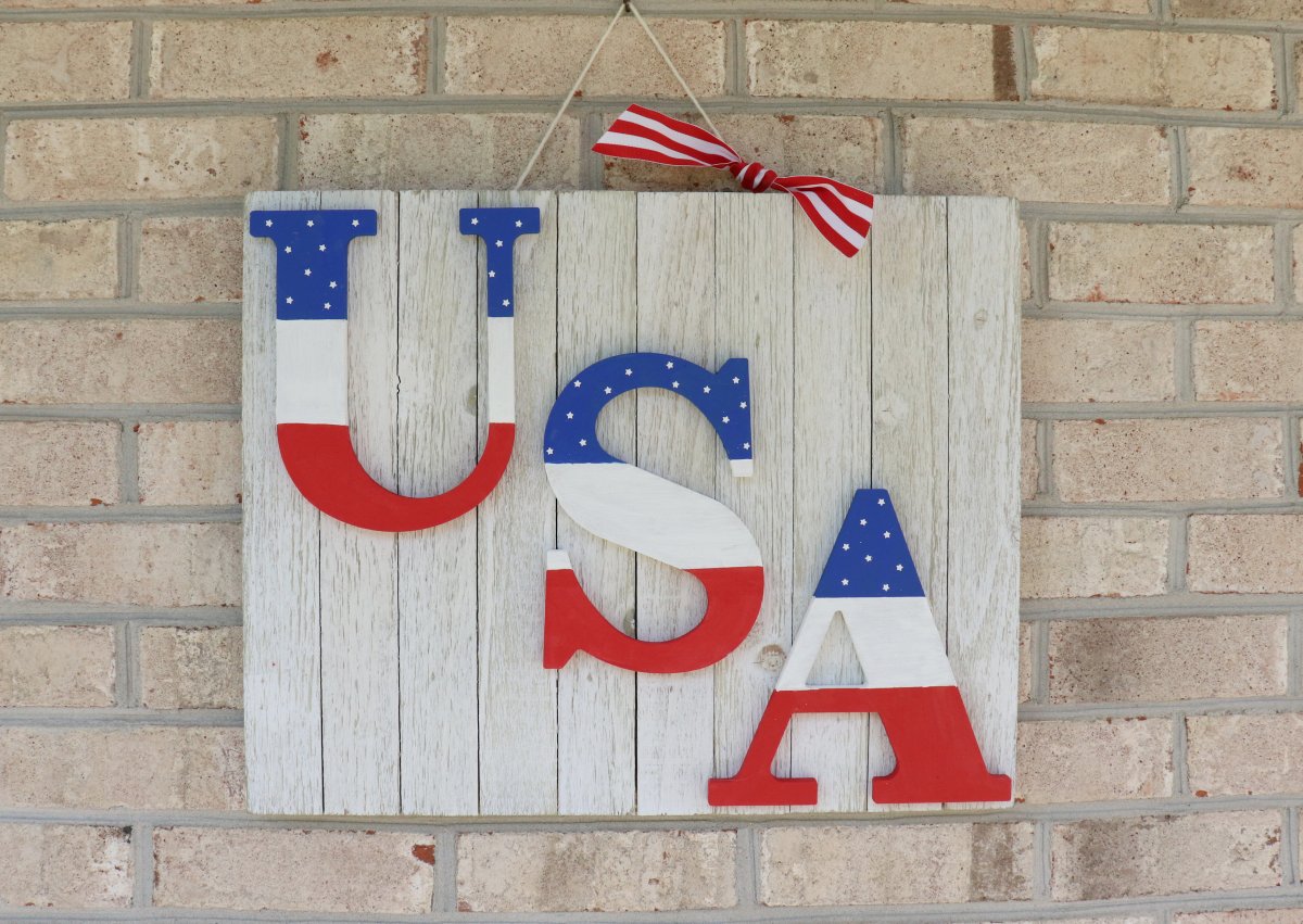 Image contains a white wood sign with the letters "USA" on it. The letters are each painted red, white, and blue. The sign hangs on a brick wall from a twine string adorned with a red and white ribbon bow.