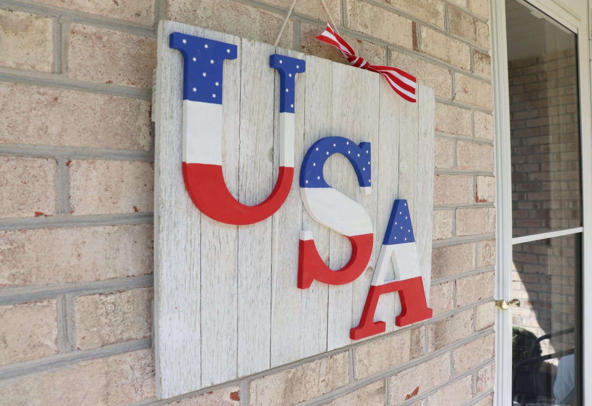 Image contains a white wooden sign with red, white, and blue "USA" letters. It hangs on a brick wall.